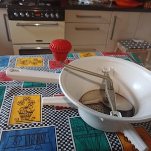 Könnte beinhalten: Eine weiße Lebensmittelmühle mit einem roten Kurbelgriff. Die Mühle hat ein Metallmesser und eine weiße Schüssel. Die Mühle steht auf einer bunten karierten Tischdecke.