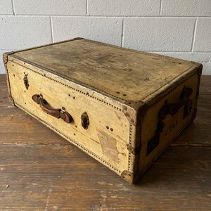 May include: A vintage, rectangular, tan-colored suitcase with a leather handle and brass hardware. The suitcase has a worn, distressed look and is made of a sturdy material.