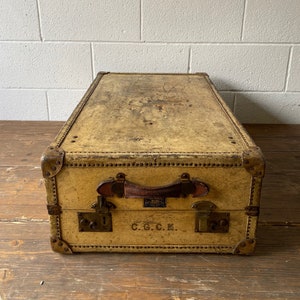May include: A vintage, tan leather suitcase with brass hardware, a leather handle, and the initials "C.G.C.K." embossed on the front.