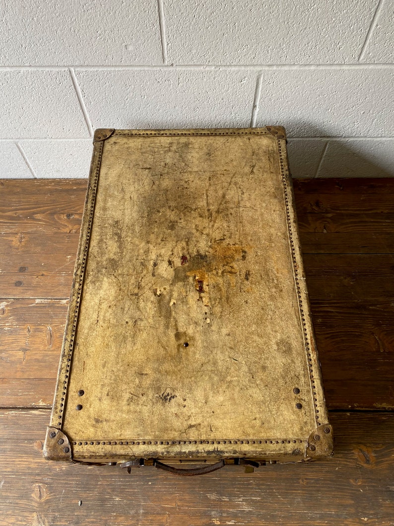 May include: A vintage leather suitcase with a worn, cream-colored exterior. The suitcase has brass hardware and a leather handle.