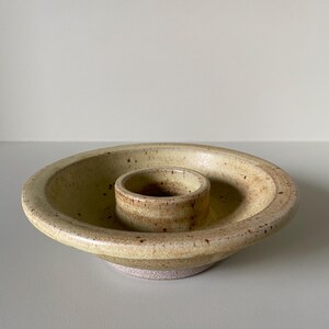 May include: A ceramic candle holder with a light yellow glaze and brown speckles. The holder has a central ring for a candle and a wider base.