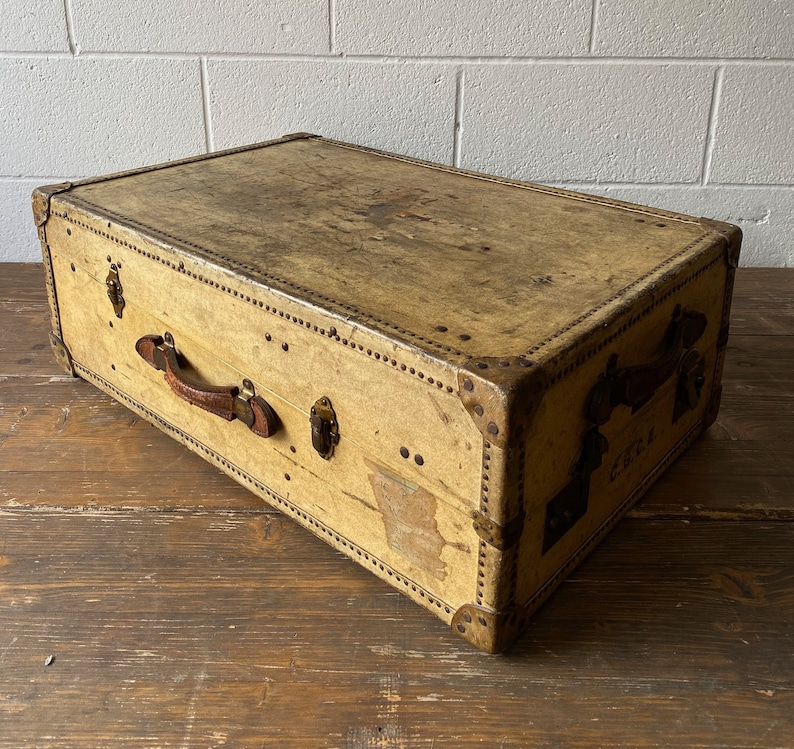 May include: A vintage, rectangular, tan-colored suitcase with a leather handle and brass hardware. The suitcase has a worn, distressed look and is made of a sturdy material.