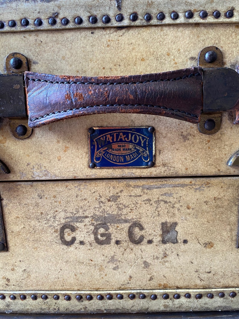 May include: A vintage, cream-colored suitcase with a brown leather handle and a blue and gold label that reads "Watajoy Regd Trade Mark London Made". The suitcase also has the initials "C.G.C.K." printed on the front.