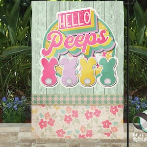 May include: A colourful garden flag with a wooden background featuring the text "Hello Peeps" in a retro font. Four pastel-coloured bunny-shaped marshmallow candies are arranged below the text. The flag has a checkered pattern and a floral pattern at the bottom.