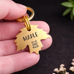 Könnte beinhalten: Ein goldfarbener, ahornblattförmiger Haustieranhänger mit dem Namen "MAPLE" in schwarzer Schrift und einer kleinen Hundeillustration. Der Anhänger ist an einem goldenen Schlüsselring befestigt.