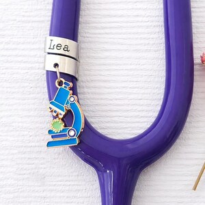 Peut inclure: Un stéthoscope violet avec une bague argentée gravée du nom "Lea". Un charm microscope bleu et doré est suspendu à la bague. Des fleurs roses séchées sont en arrière-plan.