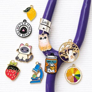 May include: A purple stethoscope with various decorative charms. Charms include a lemon, a cat, a strawberry, a microscope, and a pill bottle with cats. The charms are colorful and detailed, with a variety of shapes and designs.
