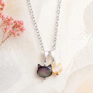 Puede incluir: Un collar de cadena plateada con dos colgantes de gato. Uno es gris oscuro y el otro amarillo claro. El collar se muestra sobre un fondo blanco y beige con flores rosas.