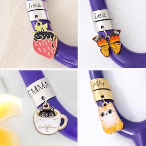 May include: Four close-up images of purple medical stethoscope tubing with personalised charms. Charms include a strawberry with a cat, a butterfly, a cat in a teacup, and a tabby cat. Each charm is attached to a band with a name.