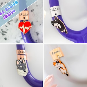 Peut inclure: Un stéthoscope violet avec des étiquettes nominatives et des breloques personnalisées. Les breloques comprennent un cœur rouge avec un battement de cœur, un chat noir et blanc, un chien husky et un chat calico. Le texte "MORE DESIGN INSIDE" est visible.