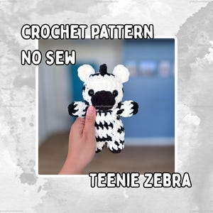May include: A small, plush, handmade zebra toy crafted from white and black yarn. The zebra features a rounded head, two small ears, and a black snout with dark, glossy eyes. The text CROCHET PATTERN NO SEW TEENIE ZEBRA is displayed in white.