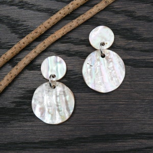 May include: A pair of iridescent white shell earrings with a round shape. The earrings are made of two pieces of shell, one smaller than the other, connected by a silver loop.