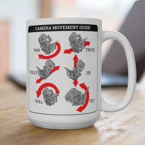 May include: White ceramic mug with a black and white illustration of a camera with red arrows showing different camera movements. The text "CAMERA MOVEMENT GUIDE" is at the top of the mug. The camera movements are labeled "PAN", "DOLLY", "ROLL", "TRUCK", "JIB", and "TILT".