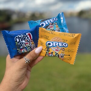 Peut inclure: Trois paquets de collations colorés sont tenus dans une main. Les paquets sont bleus et jaunes, avec les mots "Chips Ahoy!" et "OREO" imprimés dessus. Les paquets ont des illustrations de dessins animés et le texte "Gorillaz" et "Dyla".