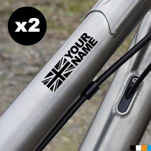 Op de afbeelding: Close-up van een fietsframe met een zwart-witte sticker met de Union Jack-vlag en de tekst "YOUR NAME". Een zwarte cirkel met "x2" staat linksboven. Zwarte kabels lopen langs het frame.
