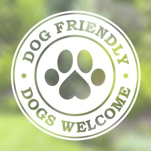 Könnte beinhalten: Ein kreisförmiges Schild mit den Worten "DOG FRIENDLY" und "DOGS WELCOME" in einem grün-weißen Design. In der Mitte befindet sich ein Pfotenabdruck-Symbol. Der Hintergrund ist ein verschwommenes Grün.
