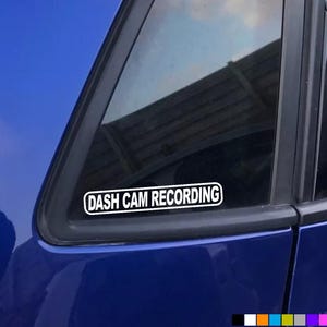 May include: A white sticker with black text that reads "DASH CAM RECORDING" is placed on the rear window of a blue car.