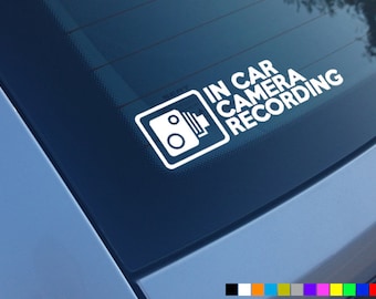 May include: White sticker with the text "IN CAR CAMERA RECORDING" and a camera icon. The sticker is on a blue car window.