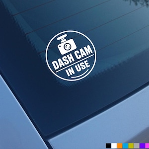 Könnte beinhalten: Ein weißer, kreisförmiger Vinyl-Aufkleber auf einer Fahrzeugscheibe. Das Design zeigt oben ein stilisiertes Kamerasymbol und darunter den Text DASH CAM IN USE in fetten Großbuchstaben, umschlossen von einer dünnen, kreisförmigen Umrandung.