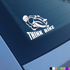 Peut inclure: Un autocollant en vinyle blanc représentant un motocycliste stylisé penché dans un virage, placé au-dessus du texte en majuscules "THINK BIKE". Le motif est présenté sous forme d'adhésif monochrome appliqué sur une surface en verre sombre.