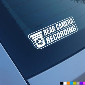 May include: A white vinyl decal applied to a car window featuring a graphic icon of a security camera and the text REAR CAMERA RECORDING in a bold, sans-serif font. The design is rectangular with rounded corners and a clean, high-contrast look.