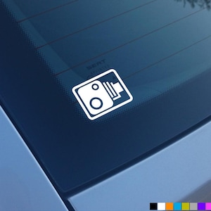 Puede incluir: Una pegatina blanca con un icono de cámara en la ventana azul de un coche. El icono es un simple contorno de una cámara con una lente y un flash.