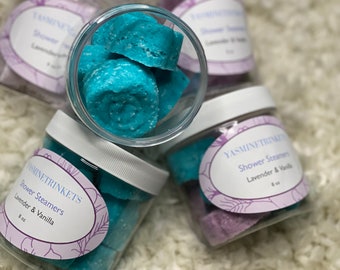 Op de afbeelding: Vier potjes met douche-stoomtabletten, twee zijn lavendel en vanille en twee zijn blauw. De potjes staan op een wit, pluche oppervlak.