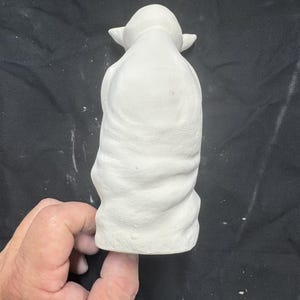 May include: A white ceramic figurine of a small creature with large pointed ears and a draped robe. The surface of the unpainted bisque pottery shows a textured fabric pattern across the body and a smooth finish on the head and ears.