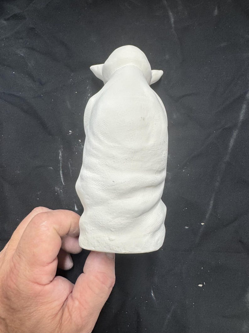 May include: A white ceramic figurine of a small creature with large pointed ears and a draped robe. The surface of the unpainted bisque pottery shows a textured fabric pattern across the body and a smooth finish on the head and ears.