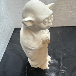 May include: An unpainted ceramic figurine of a small, elderly creature with large pointed ears, a wrinkled face, and a bald head. The figure is standing upright, wearing a long, draped robe with visible folds, and has three-toed feet.