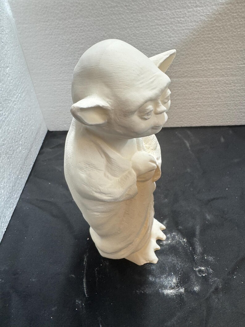 May include: An unpainted ceramic figurine of a small, elderly creature with large pointed ears, a wrinkled face, and a bald head. The figure is standing upright, wearing a long, draped robe with visible folds, and has three-toed feet.