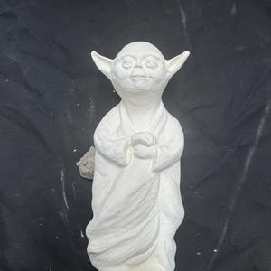 May include: A white ceramic figurine of a small, pointed-eared creature with large eyes and a wide, flat nose. The figure is depicted in a standing position, wearing a long, draped robe with visible folds, and has its hands clasped together at the chest.