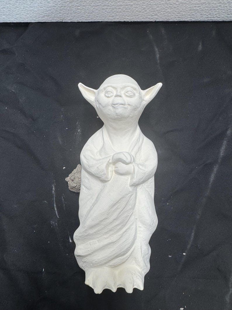 May include: A white ceramic figurine of a small, pointed-eared creature with large eyes and a wide, flat nose. The figure is depicted in a standing position, wearing a long, draped robe with visible folds, and has its hands clasped together at the chest.