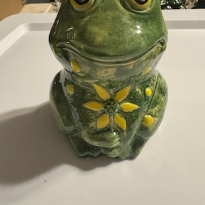 May include: A ceramic frog figurine in shades of green with yellow accents. The frog has large, expressive eyes and a sunflower design on its chest. The figurine is likely a decorative item or a container.