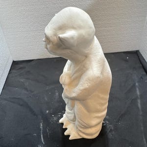 May include: A white ceramic figurine of a small, humanoid creature with large, pointed ears, a wrinkled forehead, and a robe-like garment. The figure is standing in a profile view, showing textured skin details and three-toed feet on a base.