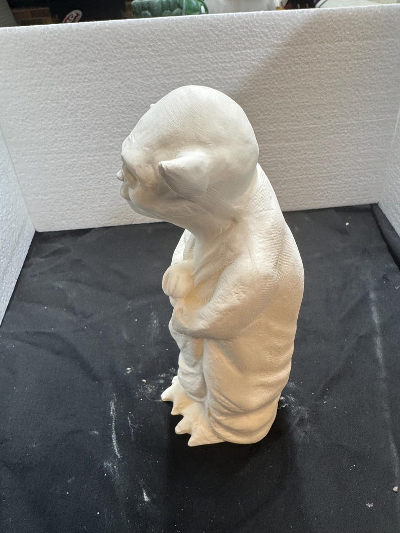 May include: A white ceramic figurine of a small, humanoid creature with large, pointed ears, a wrinkled forehead, and a robe-like garment. The figure is standing in a profile view, showing textured skin details and three-toed feet on a base.