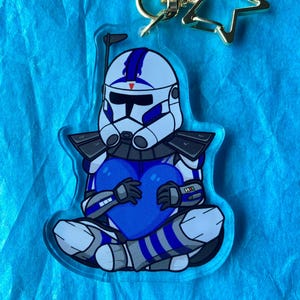 May include: A blue and white cartoon character holding a blue heart. The character is wearing a helmet and armor. The character is attached to a gold star-shaped keychain.