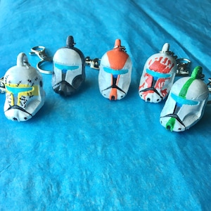 May include: Five hand-painted helmet keychains with various color schemes, including white, black, orange, and green. Each keychain features a silver keyring and a detailed design, set against a blue background.