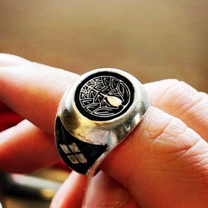 Puede incluir: Un anillo de plata con un diseño circular negro que presenta un árbol estilizado con hojas y un pájaro. El anillo se lleva en un dedo.