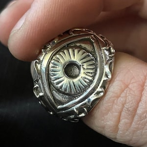 Puede incluir: Un anillo plateado con un diseño de ojo. El anillo presenta un ojo detallado, con una pupila central y líneas radiales. El anillo tiene un borde ornamentado y texturizado. El anillo está sostenido por un dedo.