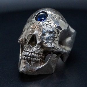 Peut inclure: Une bague en argent en forme de crâne avec une pierre précieuse bleue sertie sur le front. Le crâne est détaillé avec des sculptures complexes et une surface texturée.