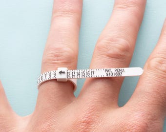 Könnte beinhalten: Ein weißer Kunststoff-Ringgrößenmesser mit einem schwarzen Pfeil und Zahlen von 6 bis 17. Der Text "PAT. PEND. 9101693" ist auf dem Größenmesser aufgedruckt.