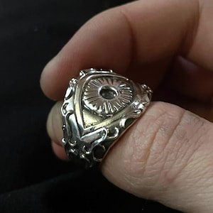 May include: A silver ring with an eye design. The eye is surrounded by intricate details and has a raised center.