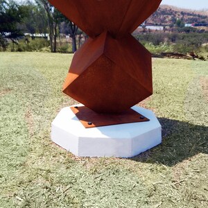 K&ouml;nnte beinhalten: Eine abstrakte Au&szlig;enskulptur mit geometrischem Design. Die Skulptur hat eine verrostete, w&uuml;rfelf&ouml;rmige Form auf einem wei&szlig;en, achteckigen Betonsockel. Die Skulptur steht auf einer Rasenfl&auml;che mit B&auml;umen und H&uuml;geln im Hintergrund.