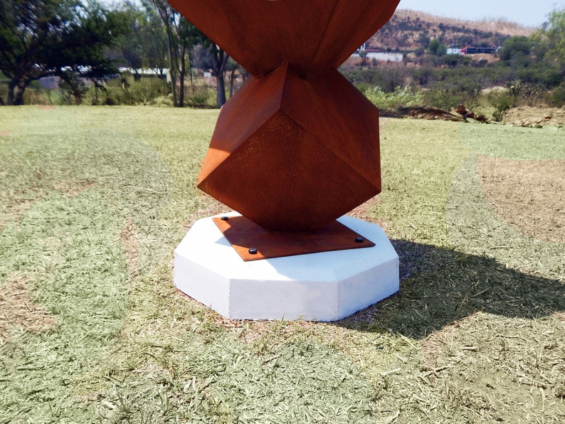 K&ouml;nnte beinhalten: Eine abstrakte Au&szlig;enskulptur mit geometrischem Design. Die Skulptur hat eine verrostete, w&uuml;rfelf&ouml;rmige Form auf einem wei&szlig;en, achteckigen Betonsockel. Die Skulptur steht auf einer Rasenfl&auml;che mit B&auml;umen und H&uuml;geln im Hintergrund.