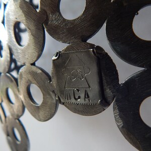 May include: Close-up of a metal sculpture made of interlocking circles. The sculpture has a small, rectangular tag attached to it with the letters "MCA" inscribed on it.