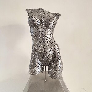 May include: A silver metal sculpture of a torso, constructed from numerous circular metal pieces. The sculpture is mounted on a rectangular, gray base. The artwork is a representation of the human form.