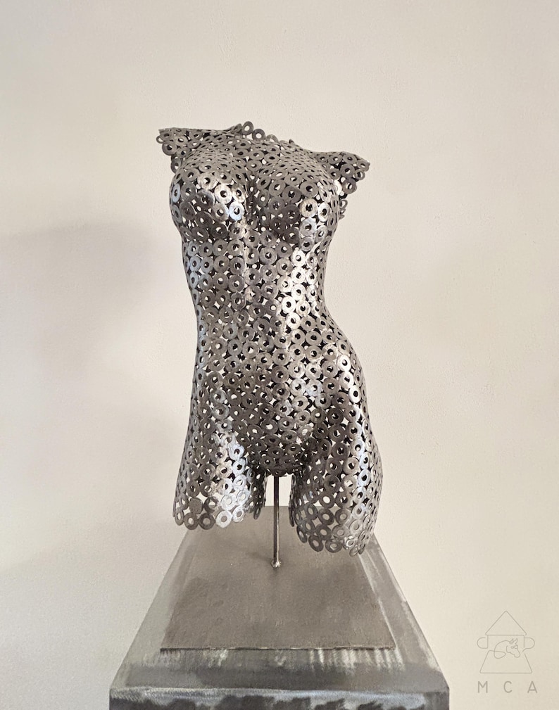 May include: A silver metal sculpture of a torso, constructed from numerous circular metal pieces. The sculpture is mounted on a rectangular, gray base. The artwork is a representation of the human form.