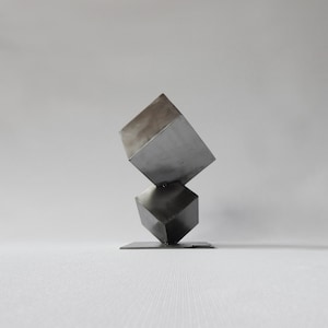 Könnte beinhalten: Eine moderne Skulptur aus zwei übereinander gestapelten Metallwürfeln. Der obere Würfel ist schräg gestellt, was ein dynamisches und abstraktes Design erzeugt.