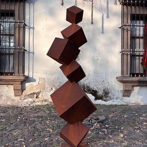 Könnte beinhalten: Eine abstrakte Skulptur aus gestapelten, rostbraunen Metallwürfeln. Die unterschiedlich großen Würfel scheinen in einem prekären Gleichgewicht zu stehen. Die Skulptur steht im Freien vor einer weißen Wand mit Fenstern.